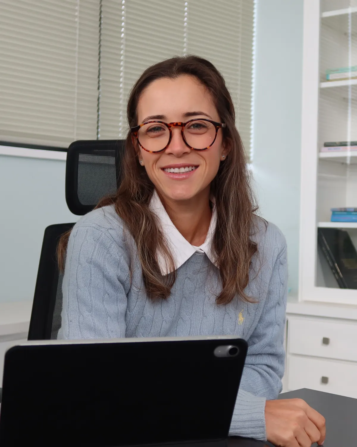 Júlia Fabris sentada à mesa do consultório, diante do tablet, sorrindo para a câmera.